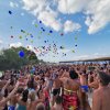 Una folla al mare lancia palloncini colorati in cielo. Giornata soleggiata con nuvole.