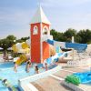 Parco acquatico con torre rossa e scivoli blu e gialli, bambini che giocano in una piscina bassa.